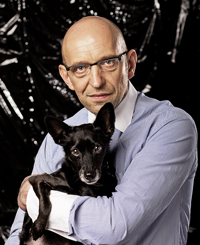 Janusz Chabior w okularach i koszuli w paski, trzyma małego czarnego psa, na ciemnym tle