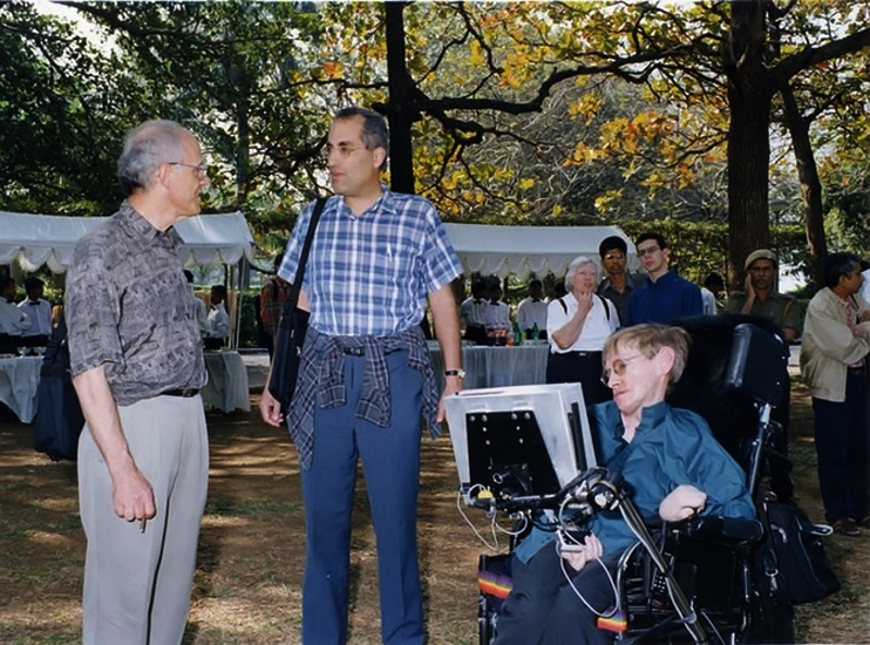 Trzech mężczyzn na zewnątrz pod drzewami: starszy siwy mężczyzna, mężczyzna w koszuli w kratę i Stephen Hawking na wózku