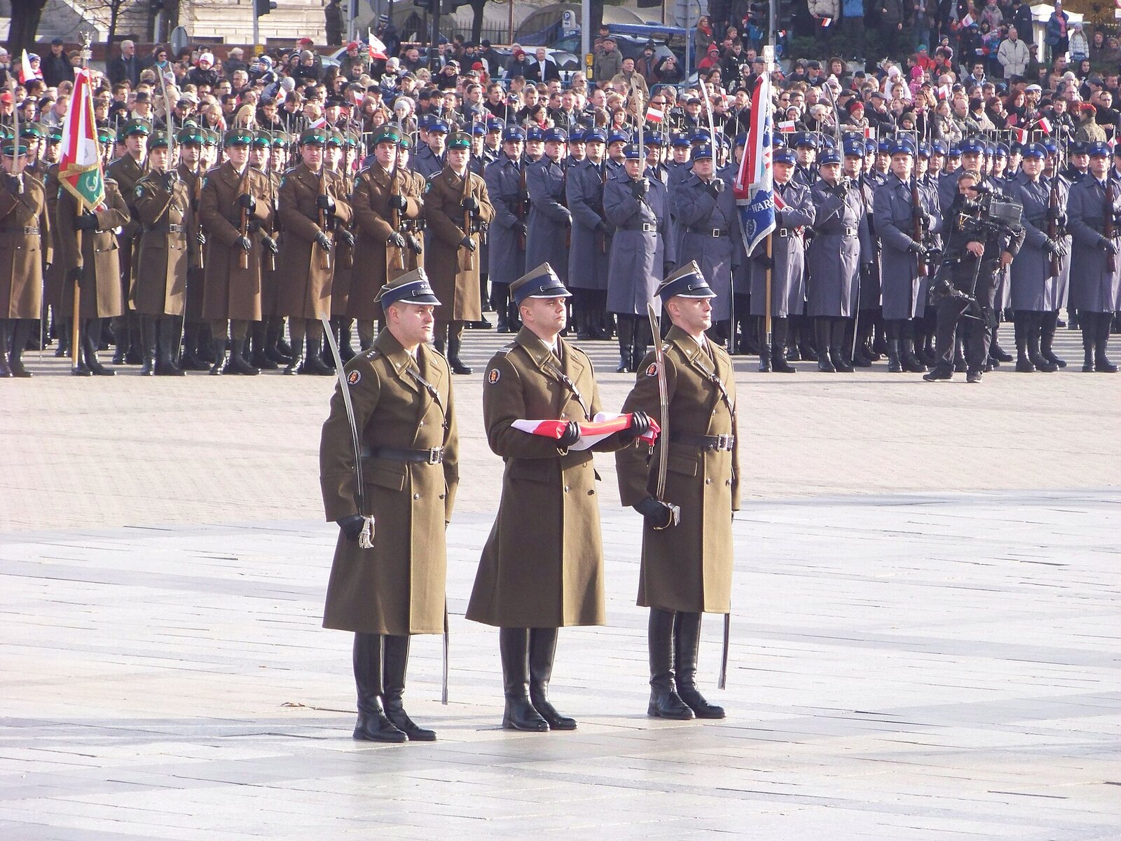 Trzech polskich żołnierzy w długich oliwkowych płaszczach, środkowy trzyma złożoną biało-czerwoną flagę; za nimi szeregi żołnierzy i tłum z chorągiewkami
