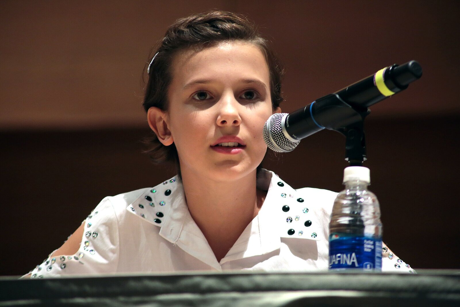 Millie Bobby Brown, dziewczyna z krótkimi ciemnymi włosami, w białej koszuli z ćwiekami, mówi do mikrofonu przy stole panelowym
